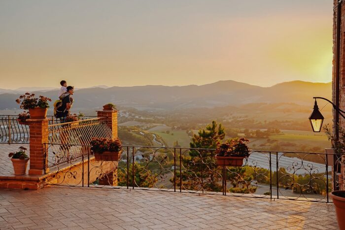 Vater mit Kind auf den Schultern blickt bei Sonnenuntergang von Terrasse auf Tal und Hügel
