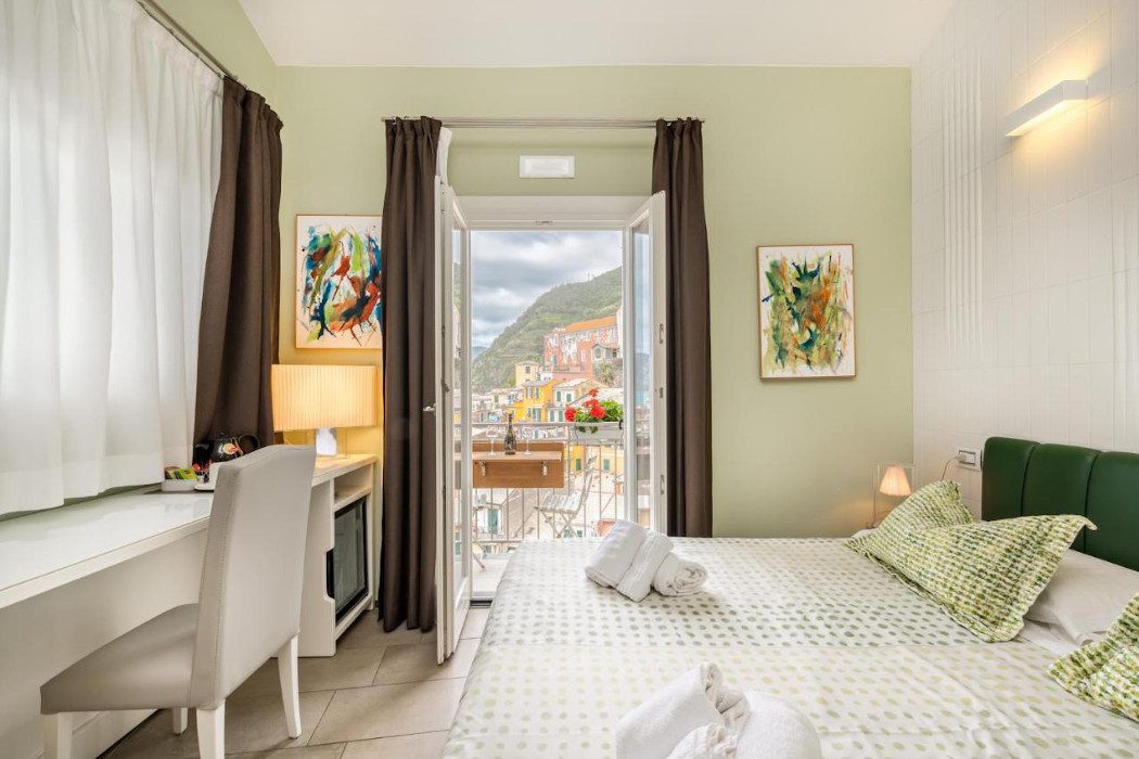 Zimmer mit Doppelbett mit grünen Kissen, Schreibtisch und Balkon mit Häusern im Hintergrund