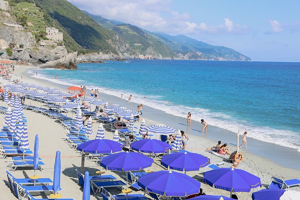 Strand mit blauen Sonnenschirmen, Menschen beim Baden, Berglandschaft im Hintergrund