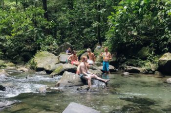 Junge Männer sitzen auf Steinen im Wasser beim La Fortuna Wasserfall in Costa Rica