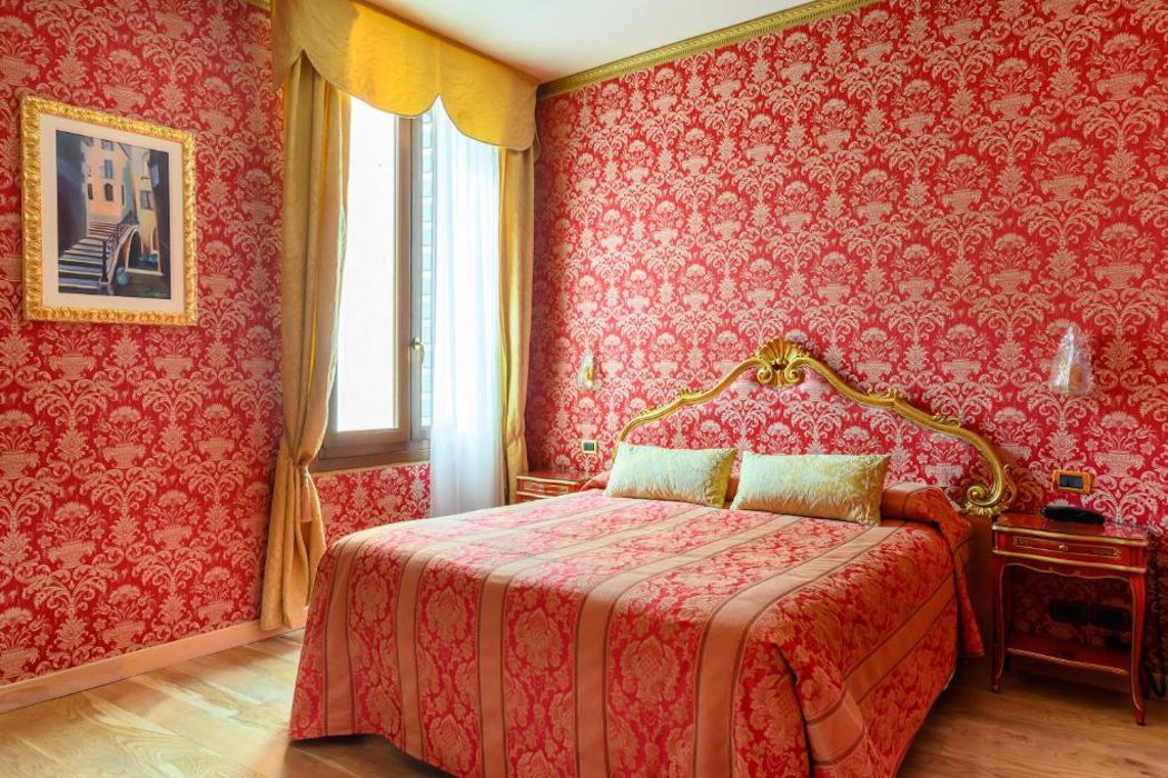 Hotelzimmer mit roter Tapete mit goldenem Muster, rotes Barock-Bett und hohes Fenster