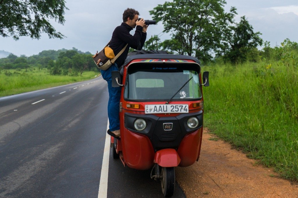 Basti auf einem kleinen, roten Auto vor einer Graslandschaft, mit der Kamera Tamron 16300 und bereit, ein Foto zu schießen